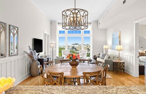 A dining area with a wooden table, chairs, a chandelier, and a view through large windows. - Designer Condo with Panoramic Gulf Views. Across from Heated Community Pool. (Inlet Beach)