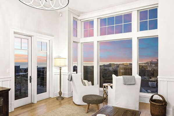 A room with large windows, a white armchair, a small stool, a wooden table, and a floor lamp.