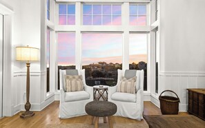 Ein Zimmer mit zwei weißen Sofas, einer Lampe und Blick auf eine Wohngegend im Sonnenuntergang.
