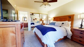 Une chambre à coucher comprenant un lit, une tête de lit en bois, des tables de chevet, un miroir, un ventilateur de plafond et une commode.