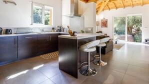 A modern kitchen with dark cabinets, a central island with bar stools, a large window, and a view of the outdoors. - Villa 4ch an idyllic haven of peace (Figari)