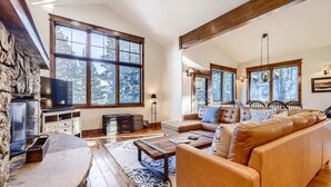 A spacious living room with a stone fireplace, large windows, a leather sofa, and a wooden dining area. - Retreat at Shock Hill Home: Walk to Gondola (Breckenridge)