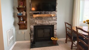 A living room with a stone fireplace, a flat-screen TV mounted above it, a wooden bookshelf, and a dining area with a table and chairs. - Sugar Ski Comfort Efficiency (with Loft) - Unit 10-303 (Sugar Mountain)