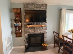 A living room with a stone fireplace, a flat-screen TV mounted above it, a wooden bookshelf, and a dining area with a table and chairs.