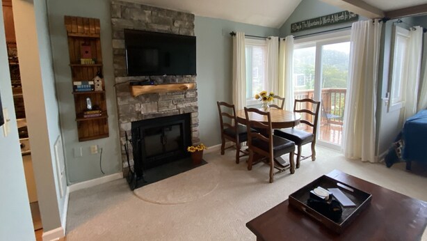 A living room with a stone fireplace, a flat-screen TV mounted above it, a dining table with chairs, and a view of the outdoors through large windows. - Sugar Ski Comfort Efficiency (with Loft) - Unit 10-303 (Sugar Mountain)