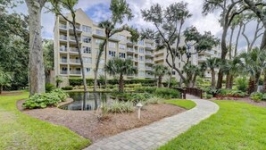 A multi-story hotel with balconies, surrounded by lush greenery and a small pond.