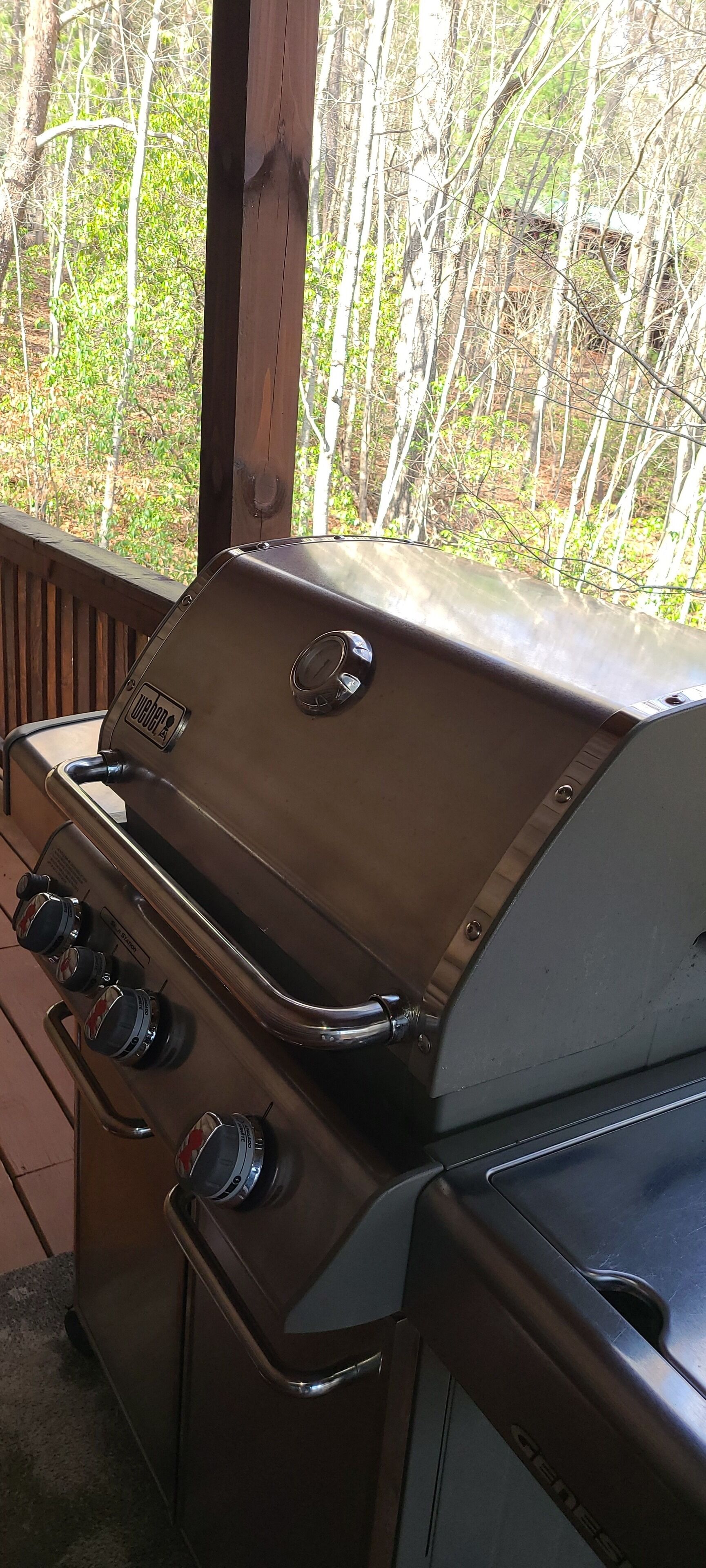 A gas grill with three burners and a control panel, located on an outdoor deck with a view of trees.