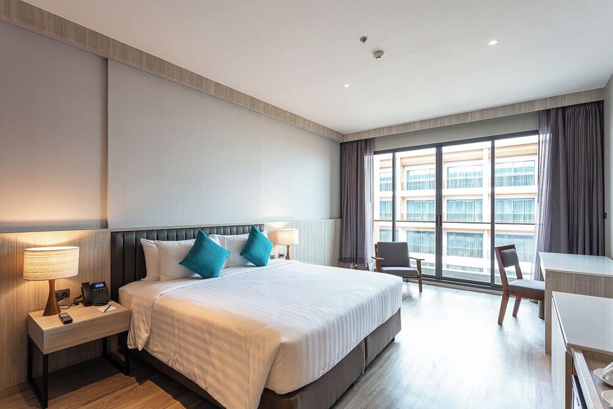 Habitación de hotel con una cama grande, mesitas de noche con lámparas, un escritorio y vistas a un edificio a través de amplios ventanales.