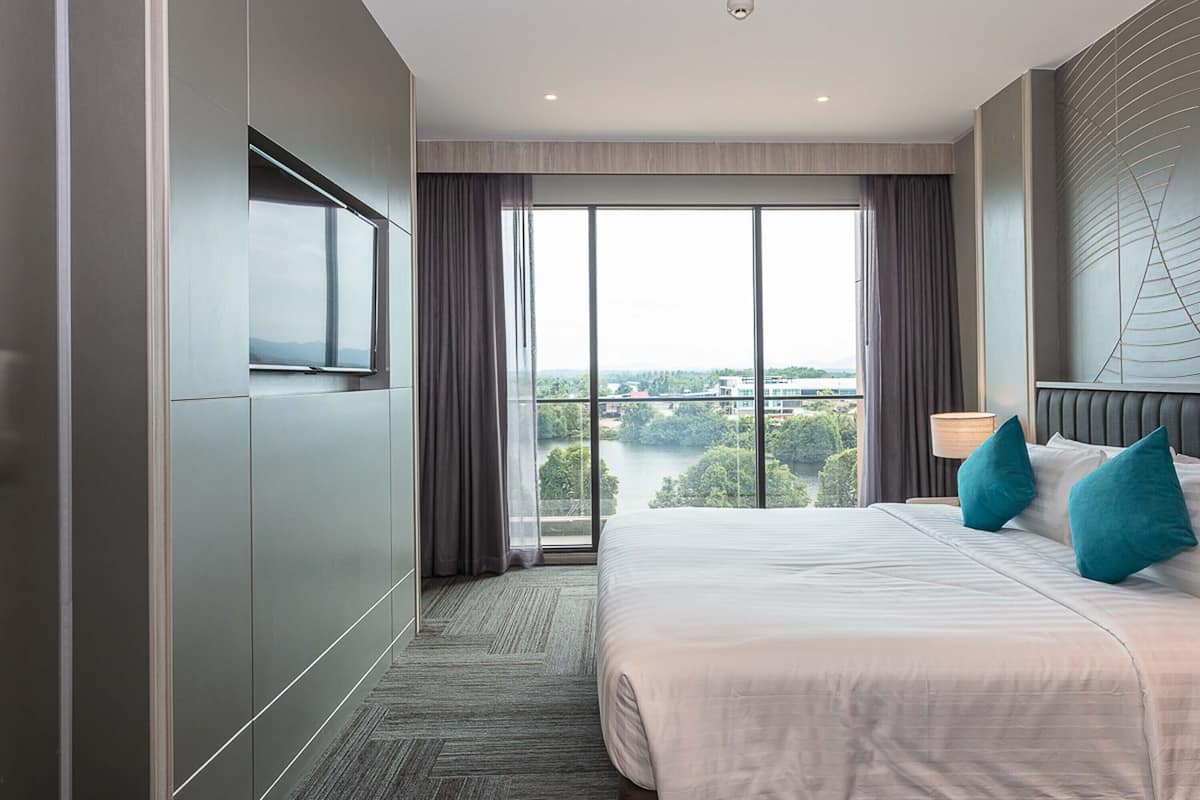 Una habitación de hotel moderna con una cama grande, televisión y vistas a un paisaje urbano a través de amplias ventanas.