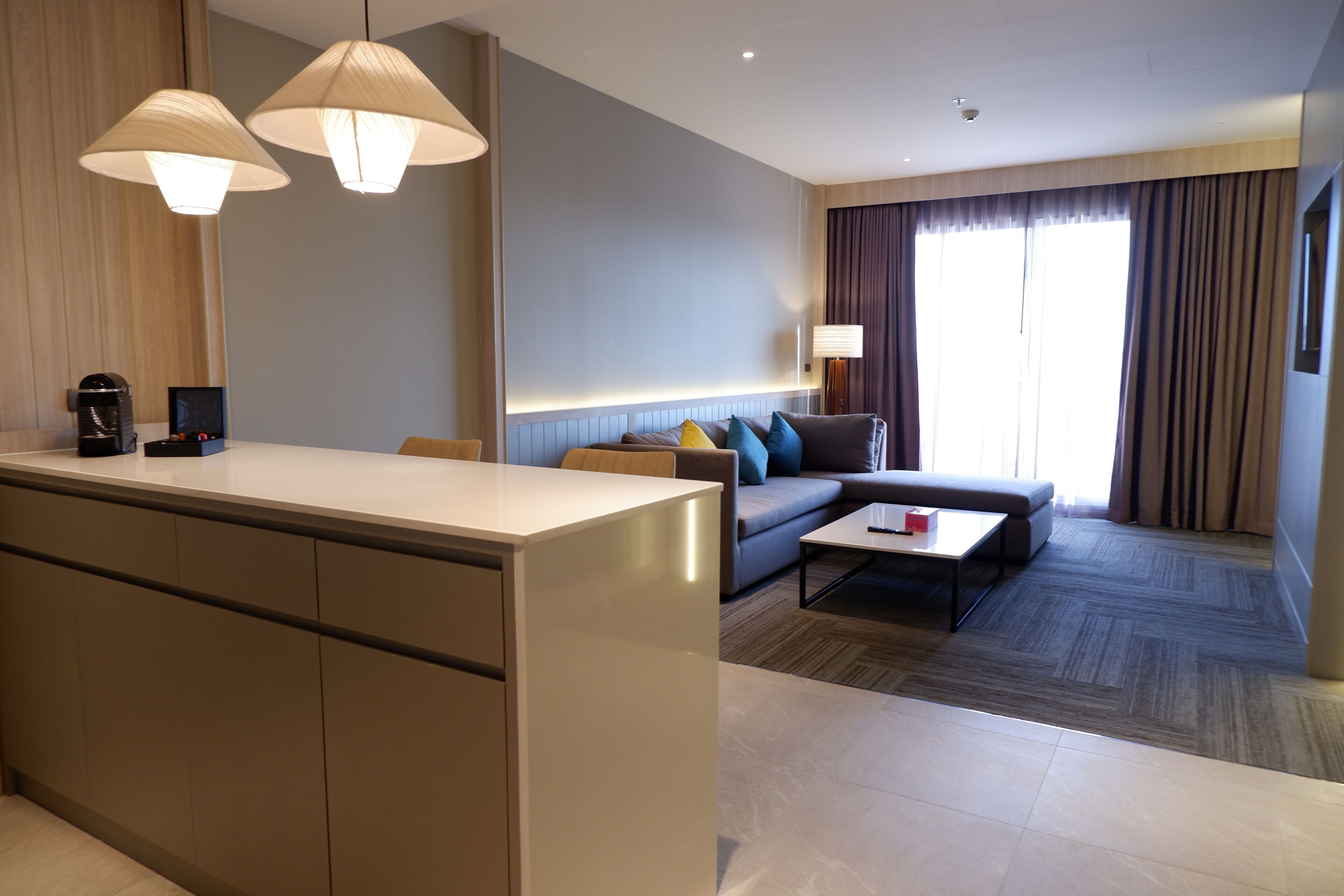 Una habitación de hotel moderna con cocina, un sofá, una mesa de centro y un ventanal con cortinas.