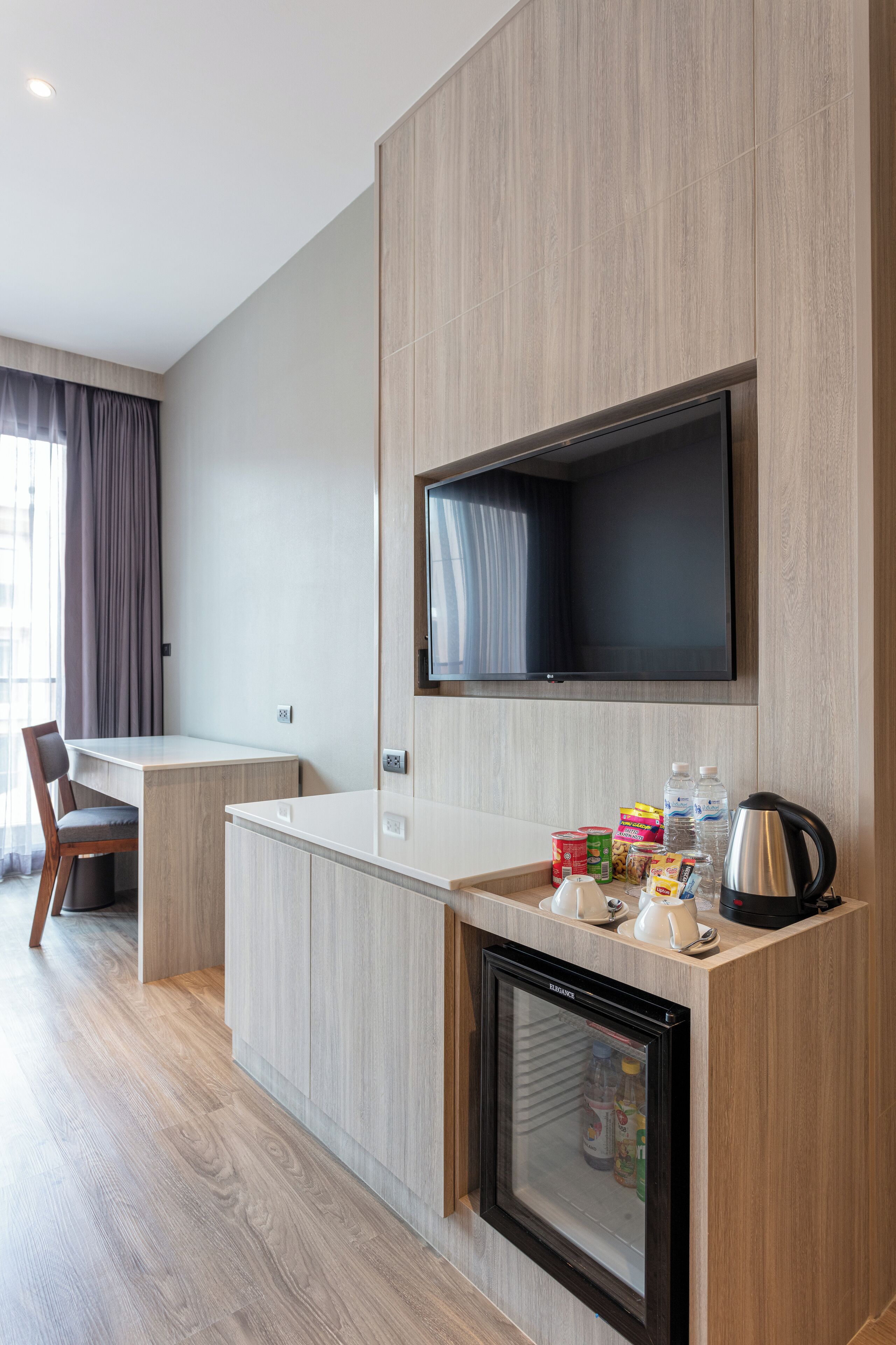 A modern hotel room with a kitchenette, a flat-screen TV mounted on the wall, a refrigerator, and a dining area with a chair and table.