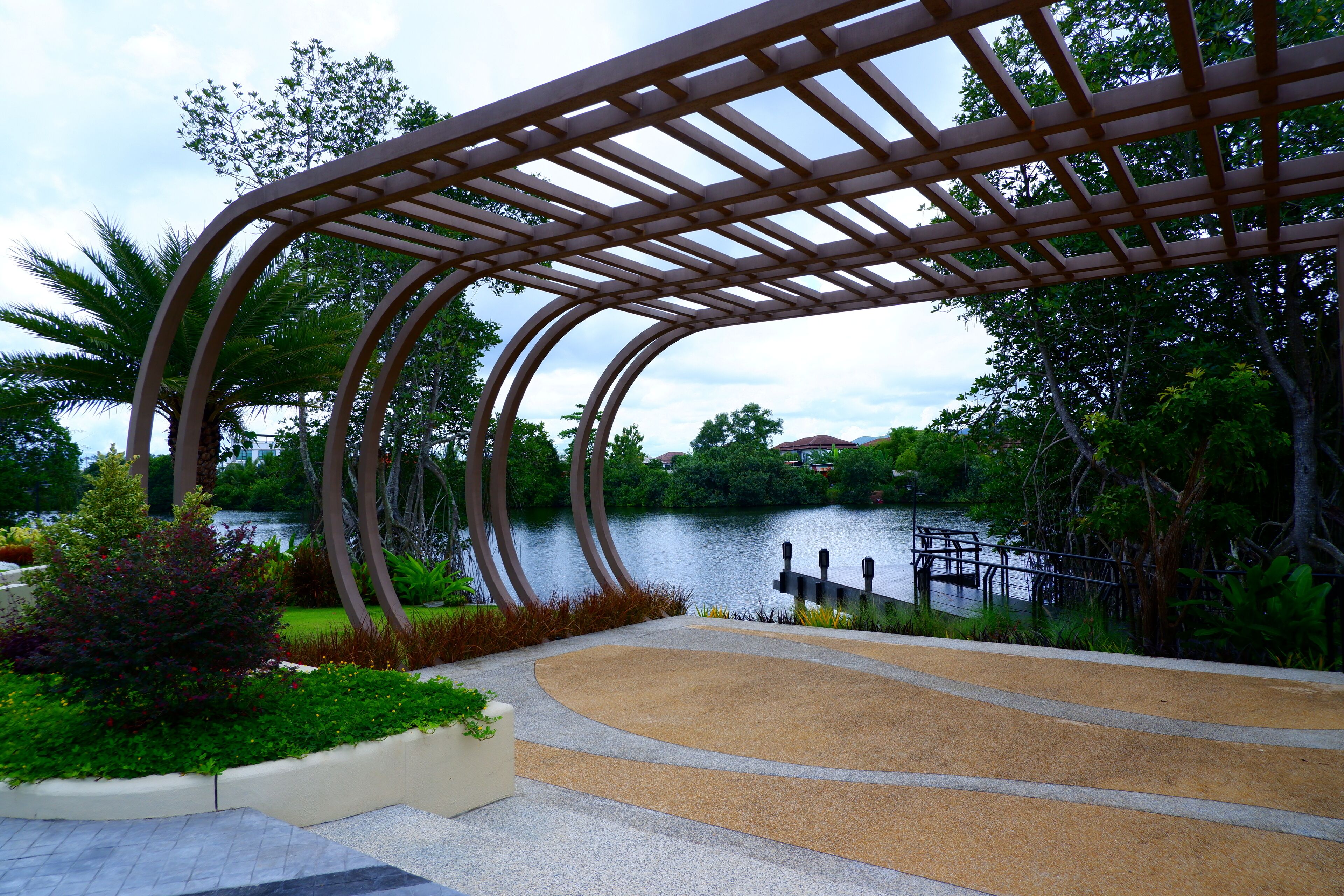 Een park met een gebogen metalen pergola, een verhard pad en een meer op de achtergrond.