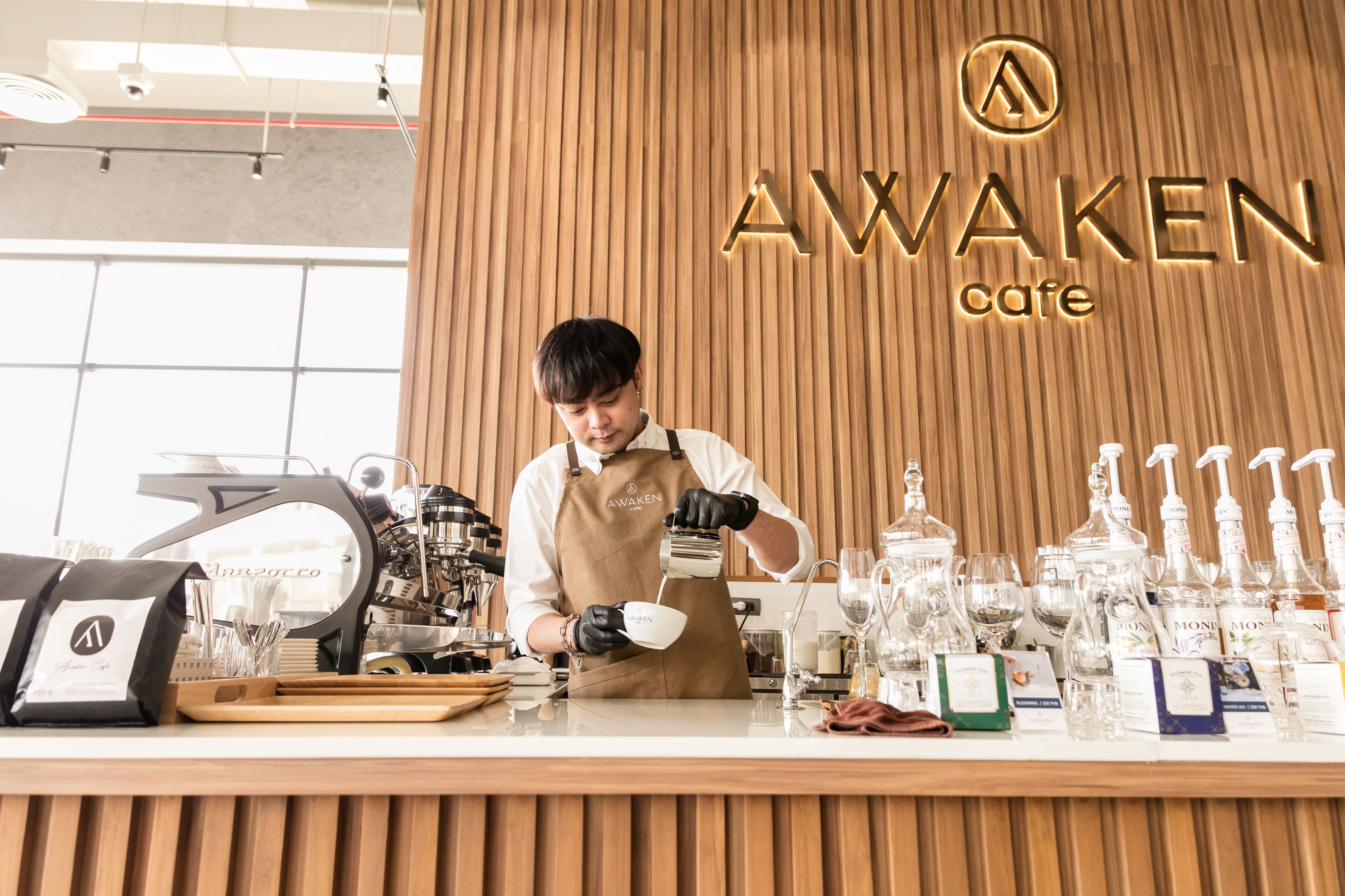 A barista at a coffee shop counter with a coffee machine, espresso cups, and various coffee beans and accessories.