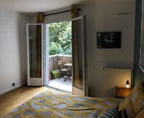 Un dormitorio con cama, una mesita, una silla y vista a un jardín por una puerta abierta.