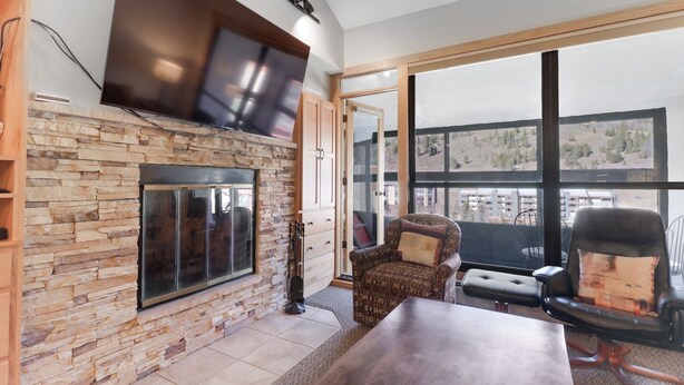 Ein Wohnzimmer mit Kamin aus Stein, einem großen, darüber montierten Fernseher und Blick auf eine gebirgige Landschaft durch große Fenster.