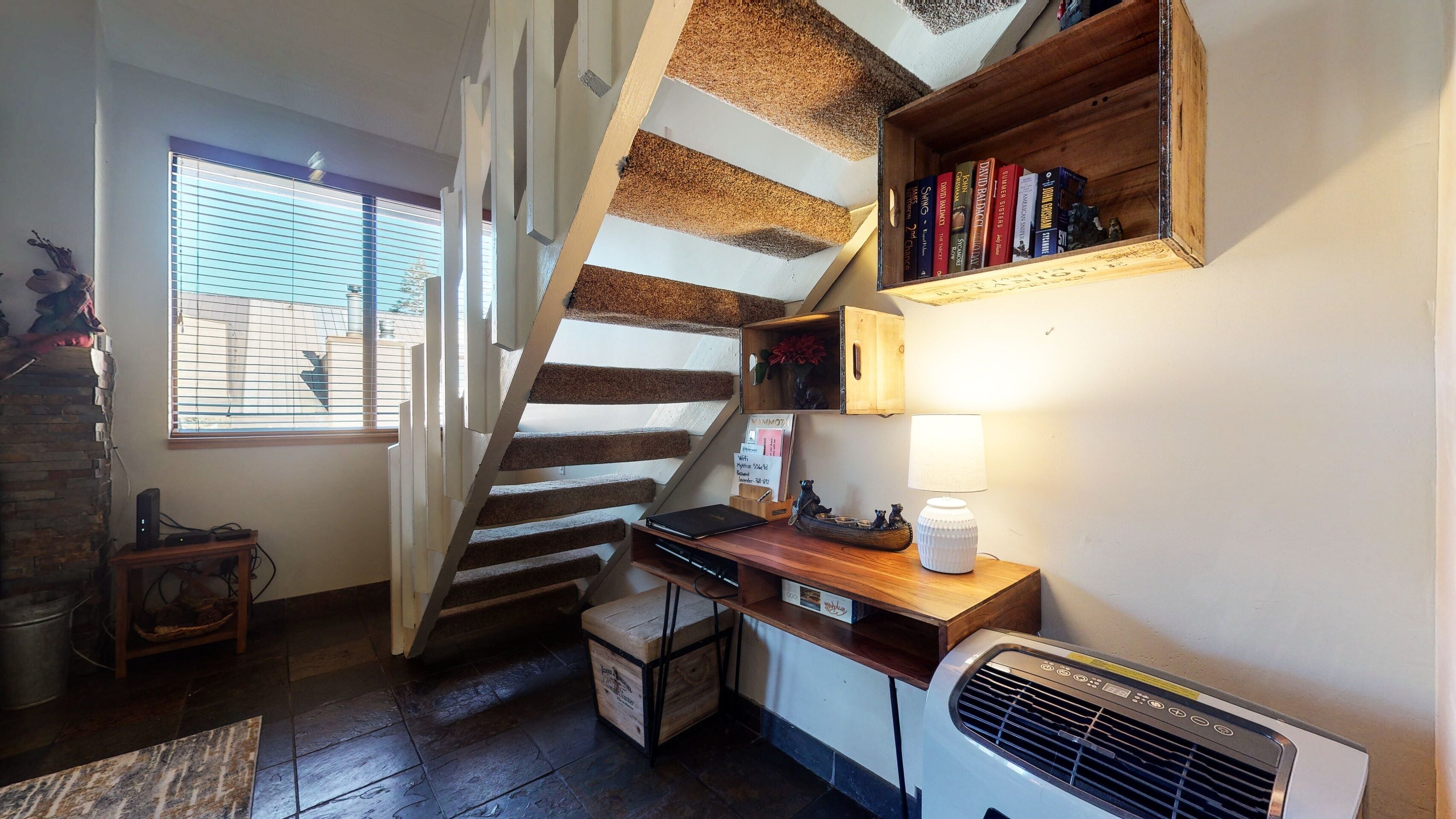 A cozy kitchen with a wooden staircase, a small table with a lamp, and a bookshelf with books.