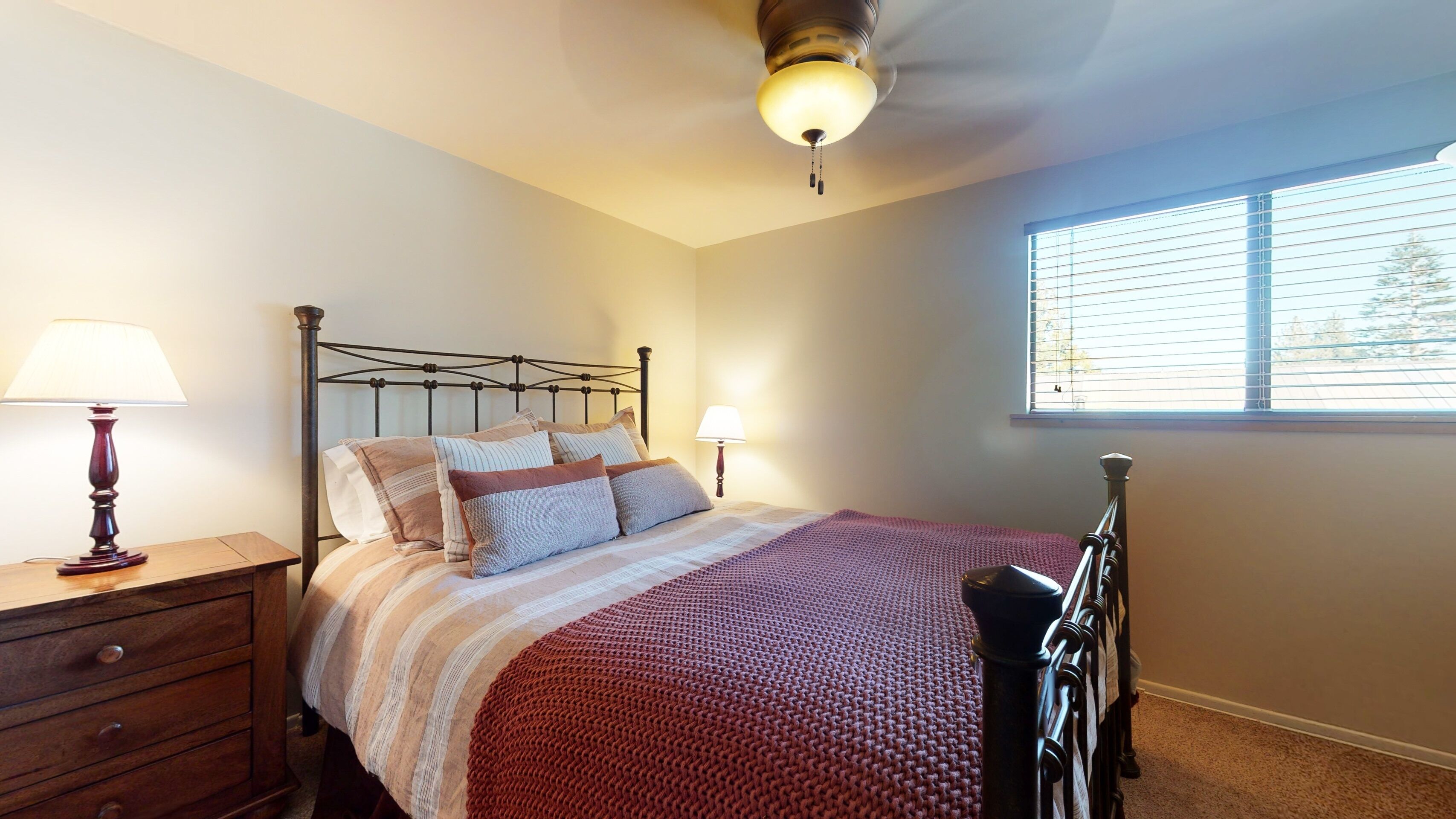 A bedroom with a bed, two bedside lamps, a wooden dresser, and a window with blinds.