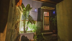 Un stretto vicolo con una piccola finestra e una porta, circondato da piante e un muro in pietra.
