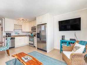A compact kitchen with white cabinets, a stainless steel refrigerator, and a microwave. There is a dining area with a wooden table and a wicker chair. A flat-screen TV is mounted on the wall, and a blue cabinet is visible.