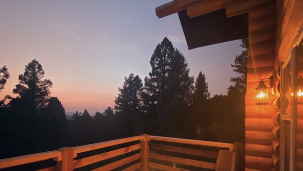 Eine Holzterrasse mit Feuerstelle, hölzernen Bänken und Blick auf Bäume im Dämmerlicht.