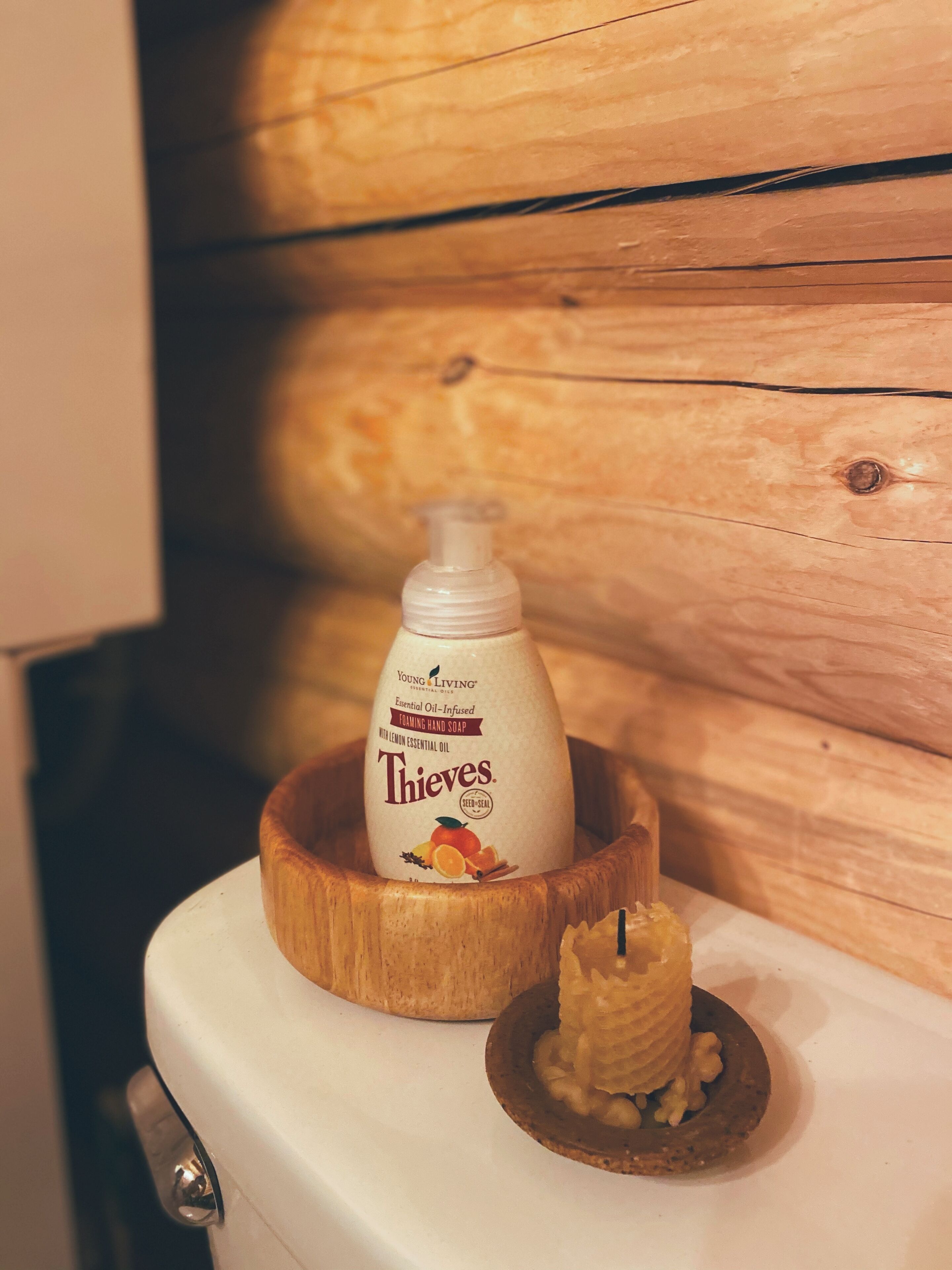 A bathroom with a wooden wall, a white toilet, a wooden bowl, a bottle of Thieves hand soap, and a candle.