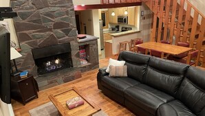 A cozy living room with a stone fireplace, a leather sofa, a wooden coffee table, and a kitchen area visible in the background. - Perfect house surrounded by National Forest to get away and decompress!!! (Winhall)