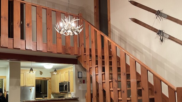 A wooden staircase with a chandelier, a dining area with a wooden table and red chairs, and a kitchen visible in the background. - Perfect house surrounded by National Forest to get away and decompress!!! (Winhall)