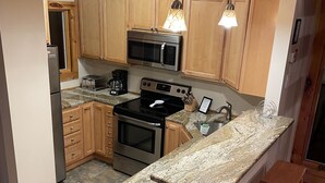 A kitchen with wooden cabinets, a stainless steel refrigerator, a microwave, an oven, a stove, and a granite countertop. There are two wooden stools and a hanging light fixture. - Perfect house surrounded by National Forest to get away and decompress!!! (Winhall)