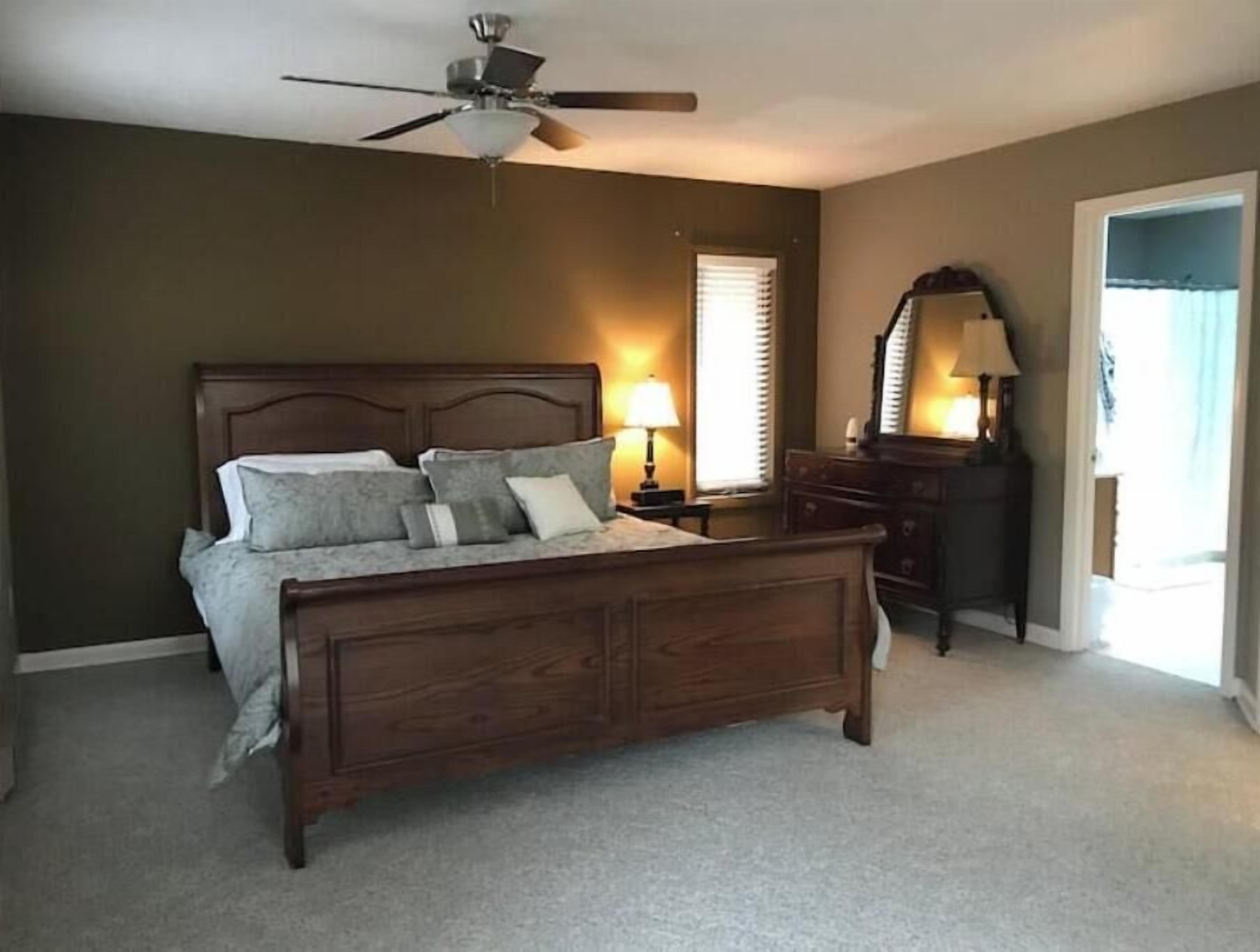 A bedroom with a wooden bed, a ceiling fan, a nightstand with a lamp, a dresser, and a mirror.