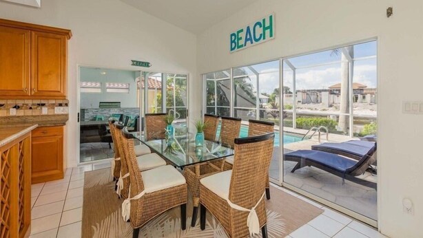 Ein Essbereich mit Glastisch und Korbstühlen, eine Küche mit Holzausstattung und Blick auf Pool und Strand durch große Fenster.