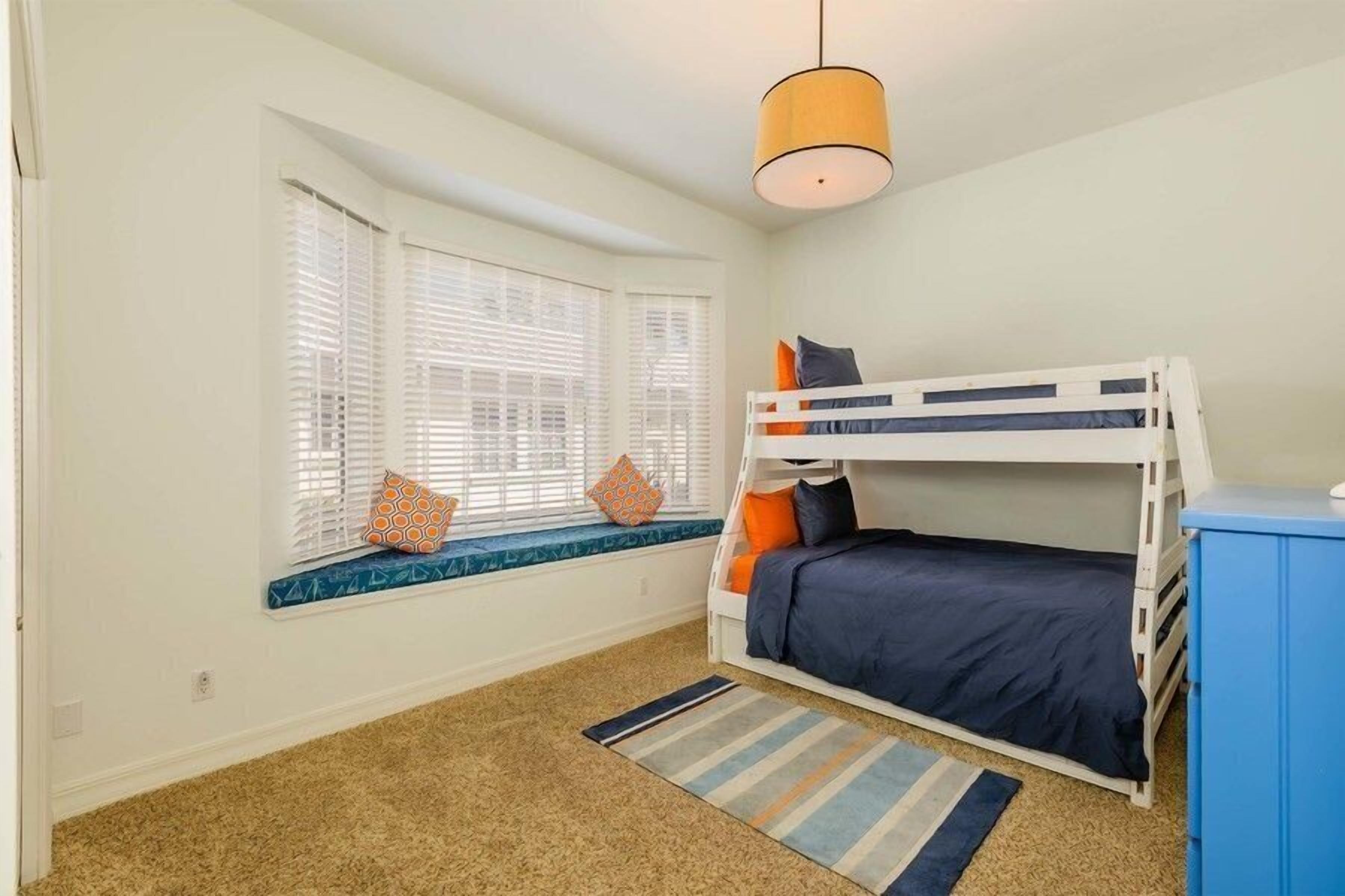 Een kamer met een stapelbed, een raam met zonwering, een blauwe kast en een gestreept vloerkleed.