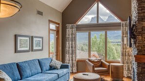 Ein modernes Wohnzimmer mit einem blauen Sofa, einem braunen Ledersessel und einem runden Hocker. An der Wand hängen zwei gerahmte Bilder, ein großes Fenster bietet Blick auf die Berge und ein Kamin aus Stein ist vorhanden.