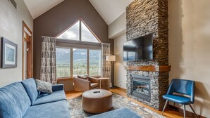 Ein modernes Wohnzimmer mit Kamin aus Stein, einem großen Fenster mit Blick auf die Berge, einem blauen Sofa und einem blauen Sessel.