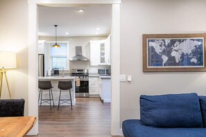 A modern kitchen with white cabinets, a stainless steel range, and a microwave. There is a dining area with a wooden table and two bar stools. A blue sofa is in the foreground, and a world map is on the wall. - Fremont Haven: Bright, Comfortable & Central (Seattle)