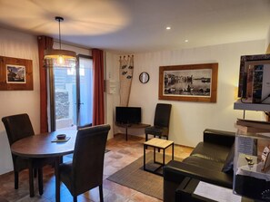 A living room with a round dining table, two chairs, a sofa, a small coffee table, and a television. There is a window with curtains, a wall-mounted clock, and a framed picture.