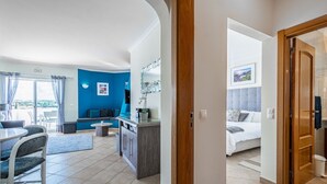 Een modern appartement met een eetgedeelte, een woonkamer met een blauwe accentmuur, een keuken en een slaapkamer met een groot bed en uitzicht op het omliggende landschap.