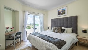 Een slaapkamer met een groot bed, een bureau, een stoel, een raam met gordijnen en een ingelijst kunstwerk aan de muur.