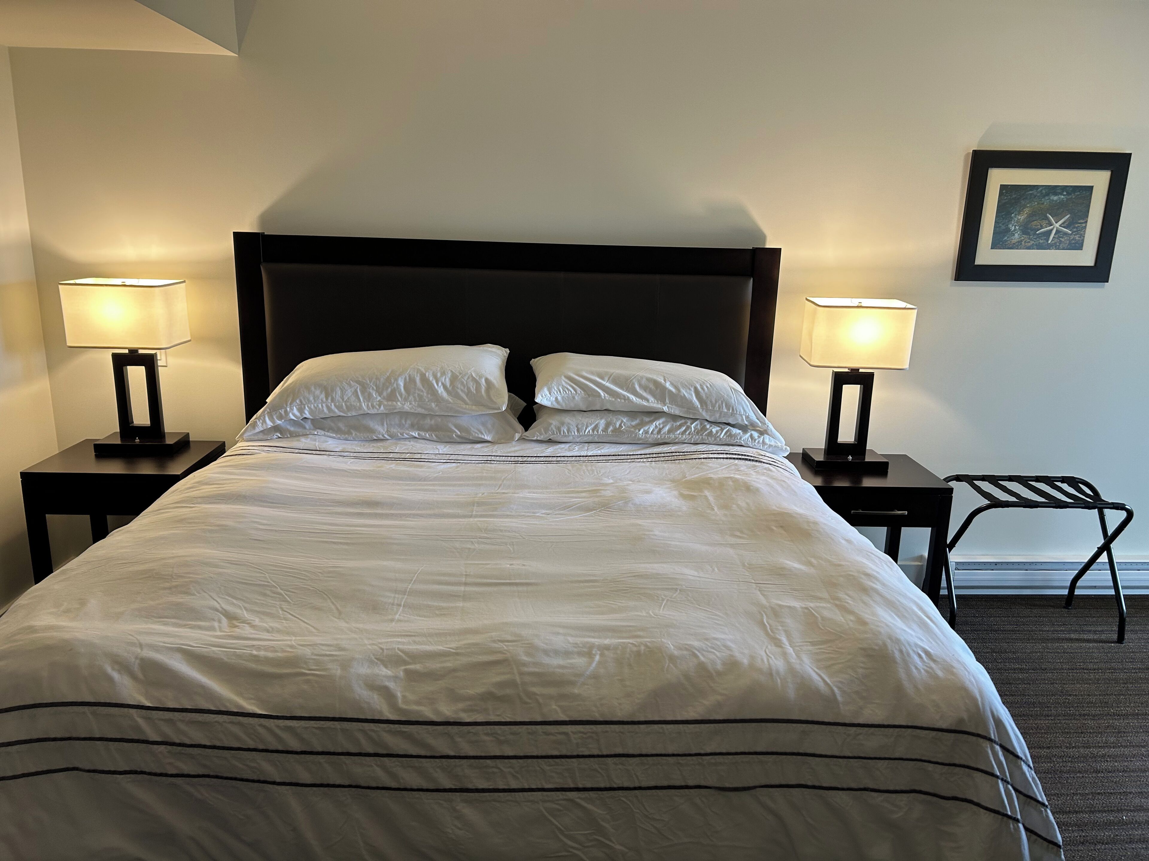 Una cama bien tendida con sábanas blancas, dos lámparas de noche, un cuadro en la pared y una silla a la derecha.