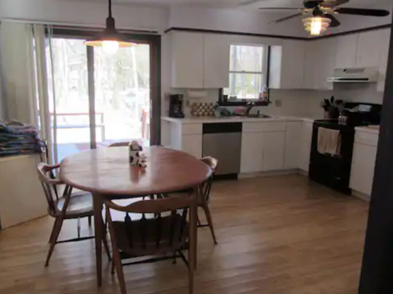 Una cucina con mobili bianchi, un tavolo da pranzo in legno con sedie, una ventola a soffitto e una finestra.