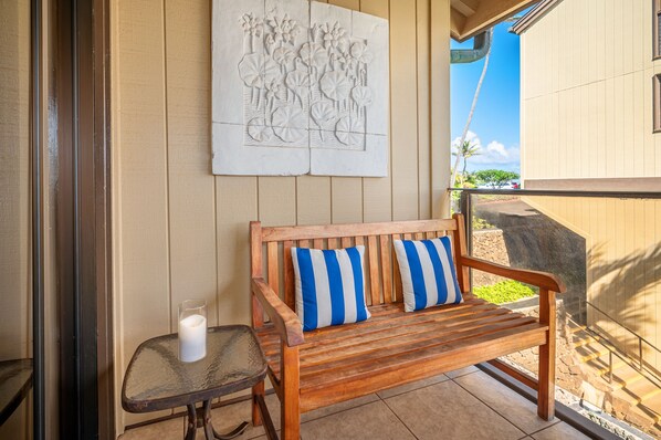 A wooden bench with blue and white striped cushions, a glass table with a candle, and a wall-mounted artwork depicting tropical flowers and birds.