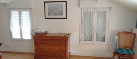 A room with a wooden dresser, a chair with a patterned cushion, and two windows with curtains.