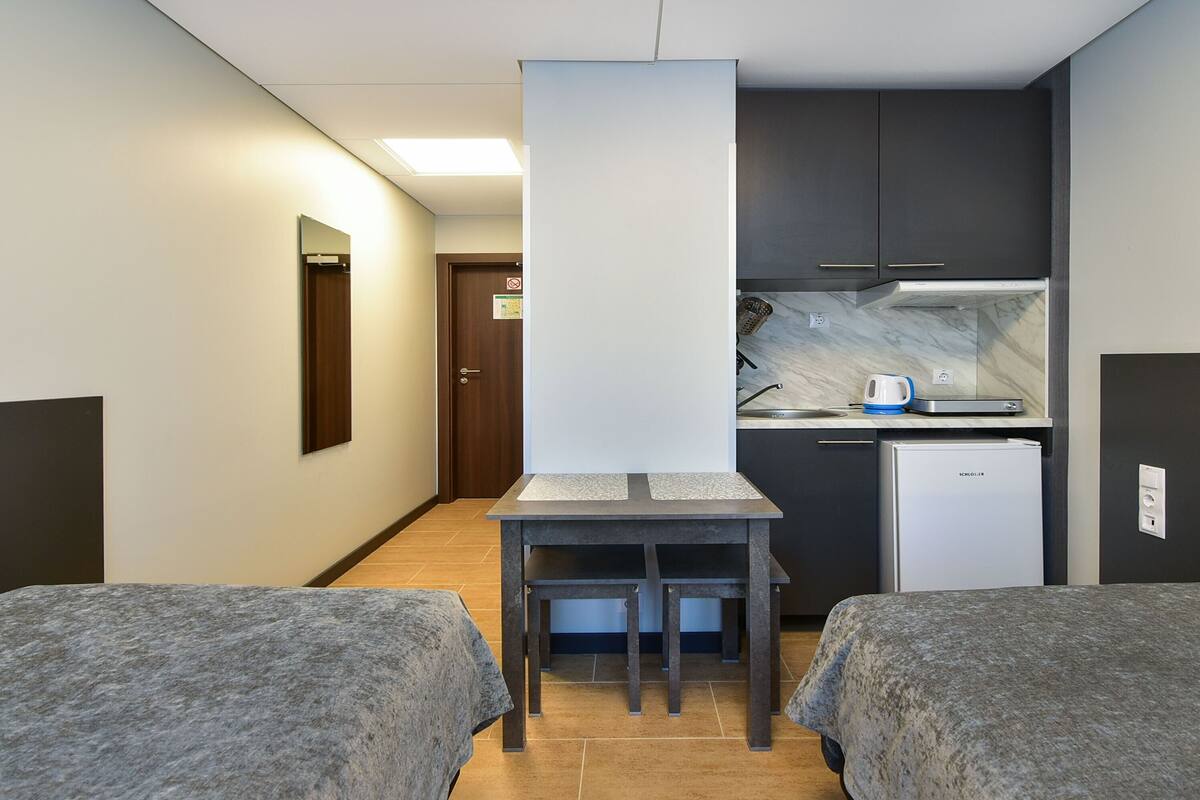 A compact hotel room with two beds, a small kitchenette, and a bathroom visible in the background.
