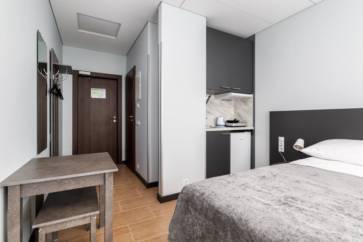 A compact hotel room with a bed, a small desk, a kitchenette, and a door leading to a hallway.