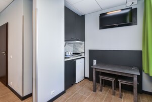 Una kitchenette compacta con un refrigerador pequeño, microondas y tetera, una mesa oscura con una silla, y un televisor de pantalla plana montado en la pared.