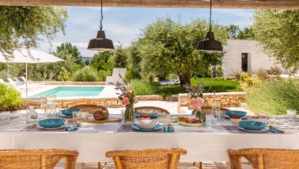 Un espacio de comedor al aire libre con sillas de mimbre, una mesa con platos y vasos azules, y lámparas colgantes.