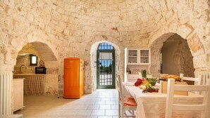 A rustic stone-walled room with a kitchen area, a dining table with chairs, and a view through an archway. - Il Trullo di Erna by BarbarHouse (Ostuni)