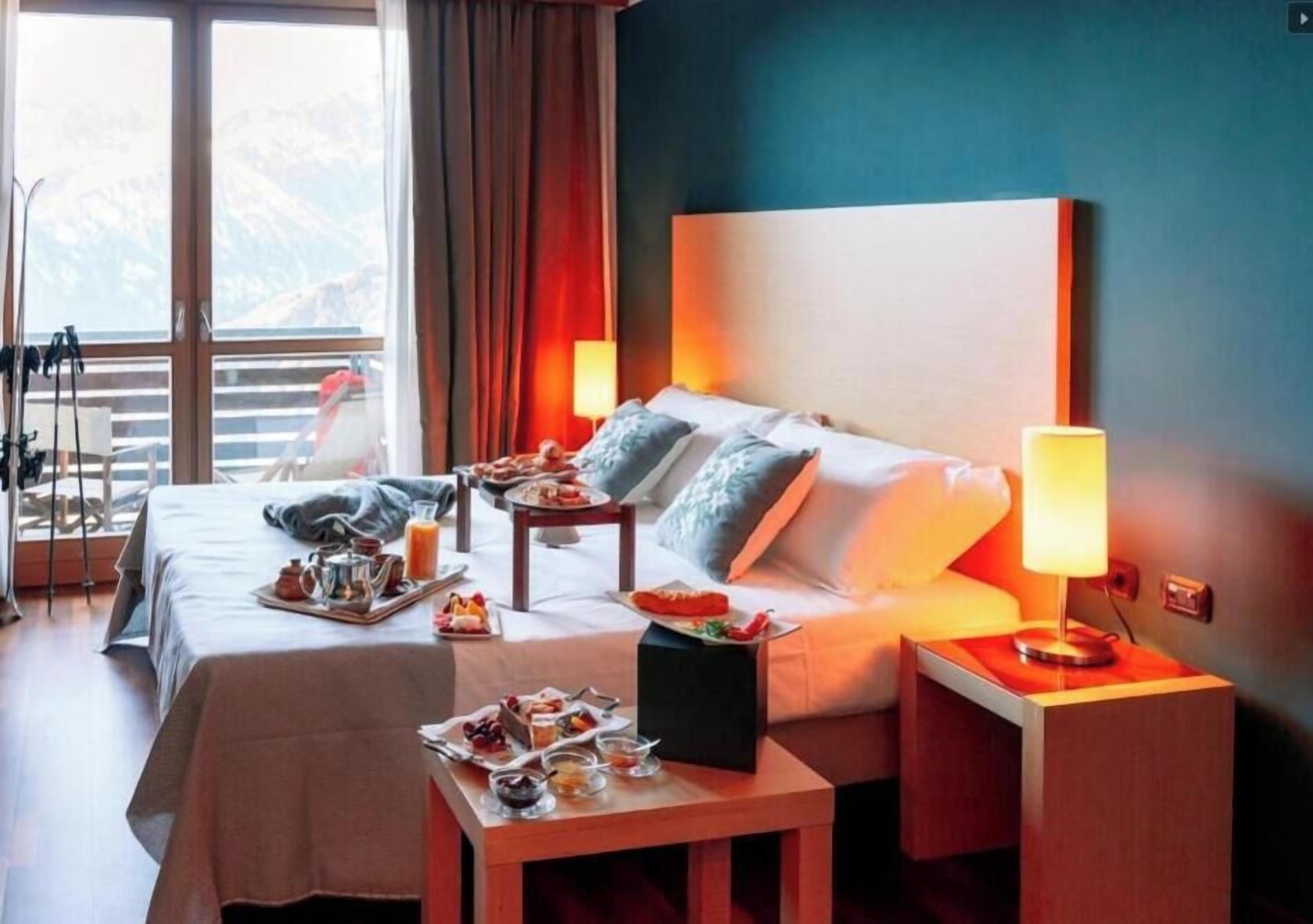 Ein Hotelzimmer mit zwei Betten, einem Esstisch mit Frühstück, einem Nachttisch mit Lampe und Blick auf Berge durch große Fenster.