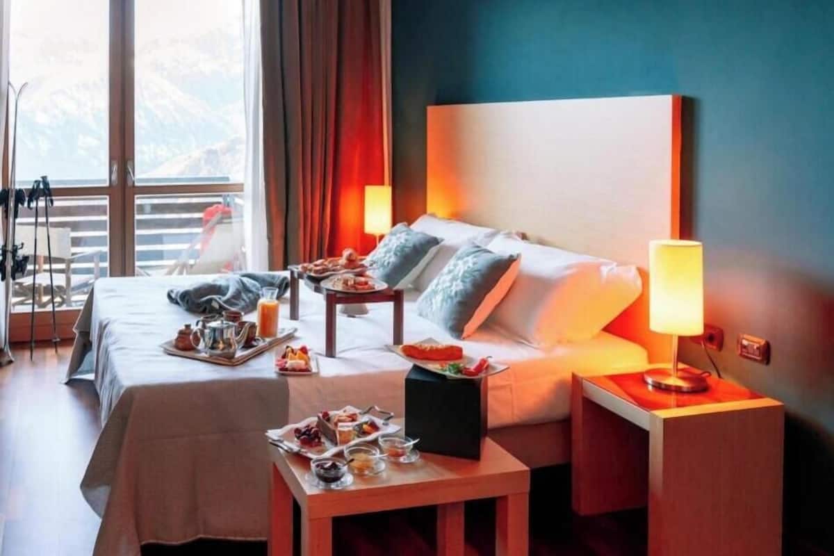 Une chambre d’hôtel avec deux lits, une table à manger où le petit-déjeuner est servi, une table de chevet avec une lampe, et une vue sur les montagnes à travers de grandes fenêtres.