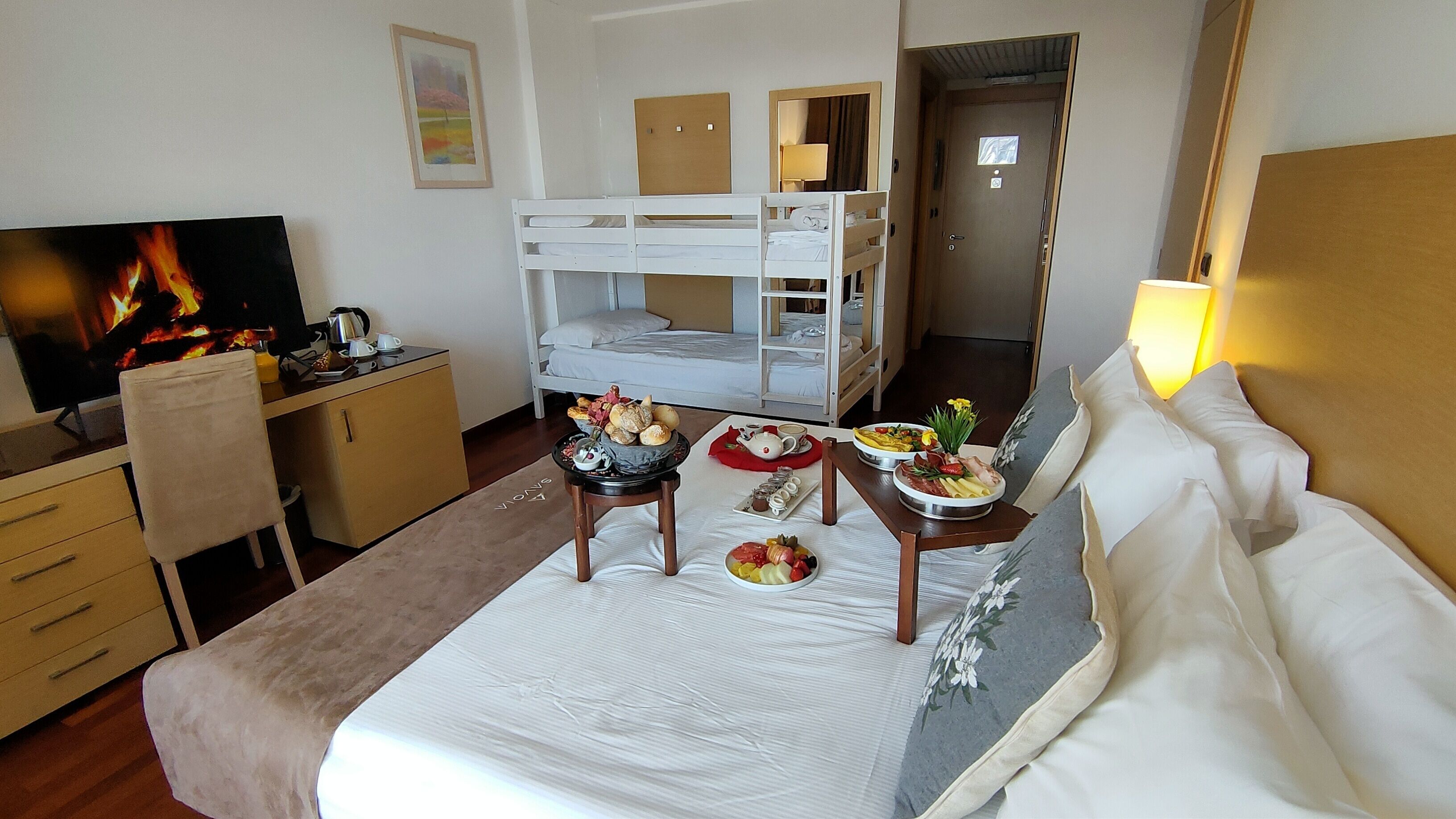 A hotel room with a bunk bed, a desk with a TV, a dining table with breakfast, and a bed with white linens.