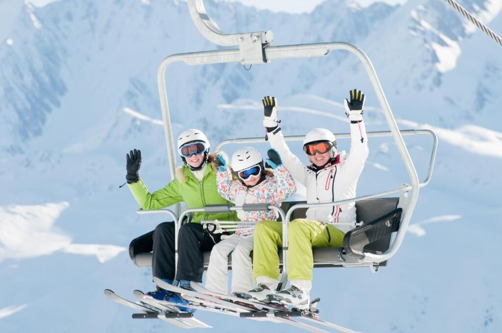 Tres personas en un telesilla con montañas nevadas al fondo.